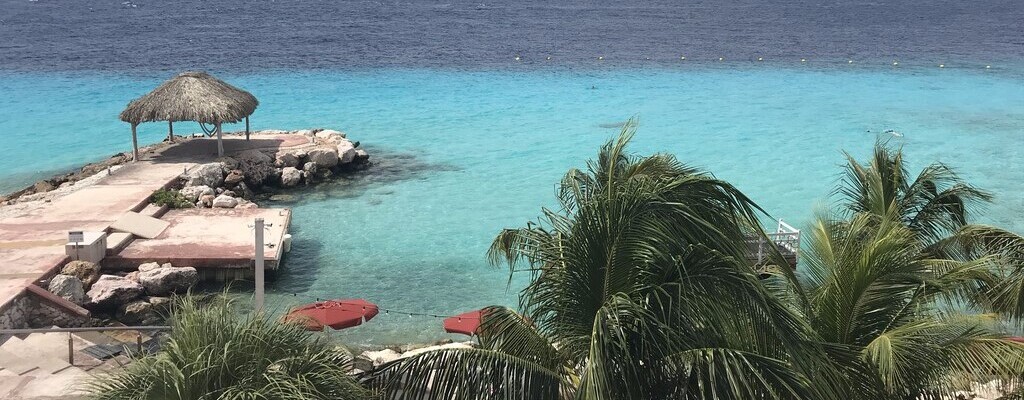 One of the ten most beautiful Curaçao beaches. You can see the Coral Estate with green palm trees in the foreground. In the background, a jetty leads into the turquoise water.