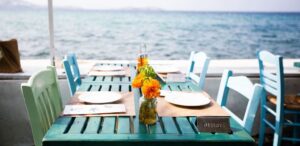 Symbolbild "Restaurant Curaçao": Ein Tisch mit vier bunten Stühlen steht auf einer Terrasse eines Restaurants. Von dort aus hat man einen hervorragenden Blick auf Meer.