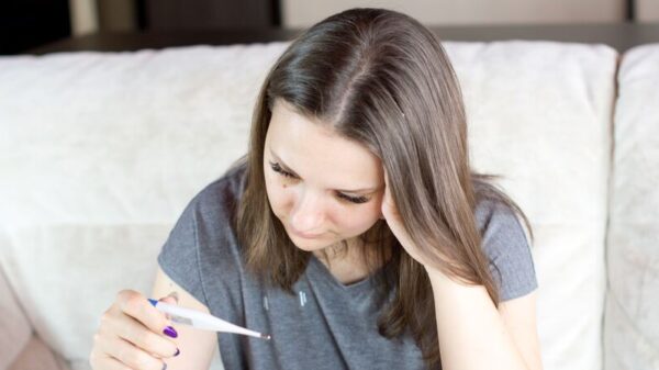 Symbolbild "Reiserücktrittsversicherung Vergleich": Eine Frau mit braunen, schulterlangen Haaren schaut betrübt auf ein Fieberthermometer.