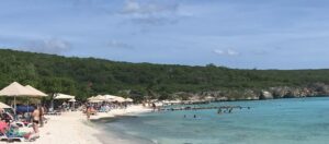 View of the sea at Playa Porto Mari in Curaçao. You can see part of the light-colored sandy beach on the left. People are lying there under parasols. On the right side of the picture you can see the blue sea.