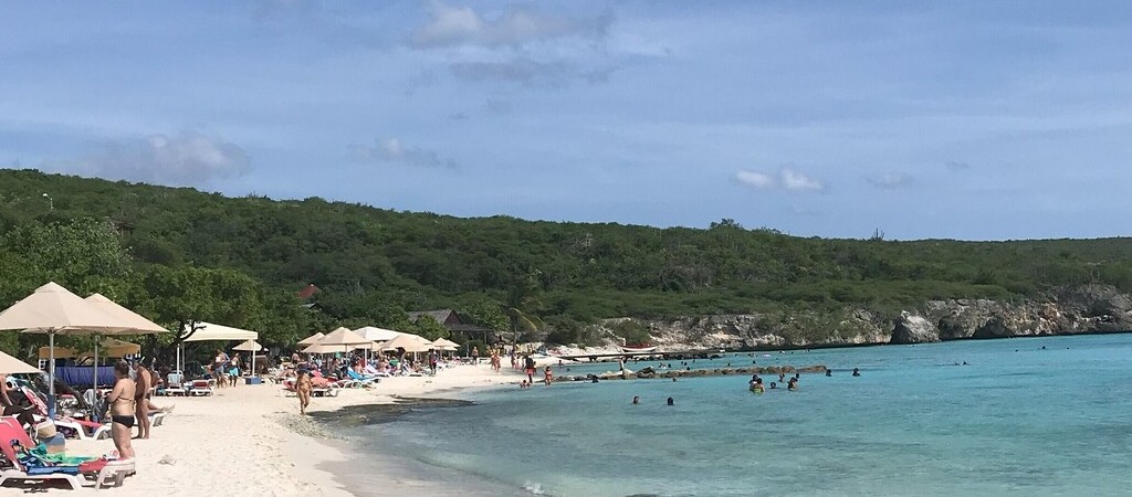 Blick auf das Meer am Playa Porto Mari auf Curaçao. Man sieht links einen Teil des hellen Sandstrandes. Dort liegen Menschen unter Sonnenschirmen. Auf der rechten Seite des Bildes sieht man das blaue Meer.