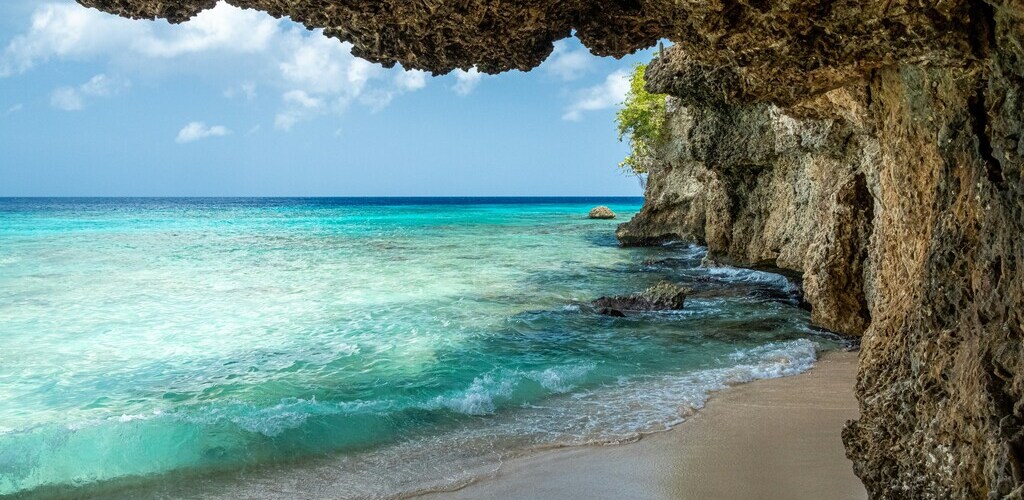 Symbolbild "ABC-Inseln Reisezeit": Zu sehen ist ein Strand auf Curaçao. Auf der rechten Seite sind Klippen zu sehen, links das türkisfarbene Meer.