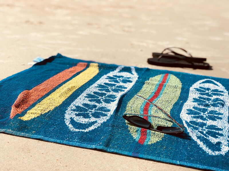 Symbolbild "Curaçao Sicherheit": Buntes Handtuch liegt am Strand. Auf dem Handtuch liegt eine schwarze Sonnenbrille. Neben dem Handtuch liegt ein Paar Flip Flops.