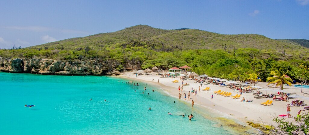 Symbolbild "ABC-Inseln": Zahlreiche Urlauber verweilen am Grote Knip, einem bekannten Strand auf Curaçao. Im Hintergrund sieht man eine grüne Landschaft, auf der linken Seite das blaue Meer.