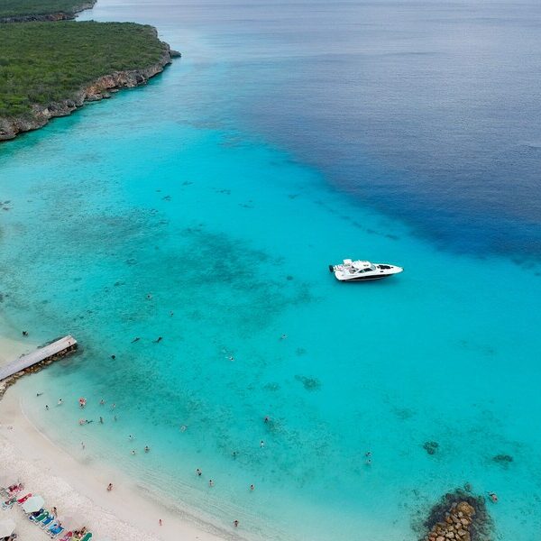 Luftaufnahme von der Küste Curaçaos, einer der Niederländischen Karibikinseln. Ein kleines Boot fährt über das tiefblaue Meer. Links sieht man einen schönen Landschaftsstreifen und einen hübschen Sandstrand.